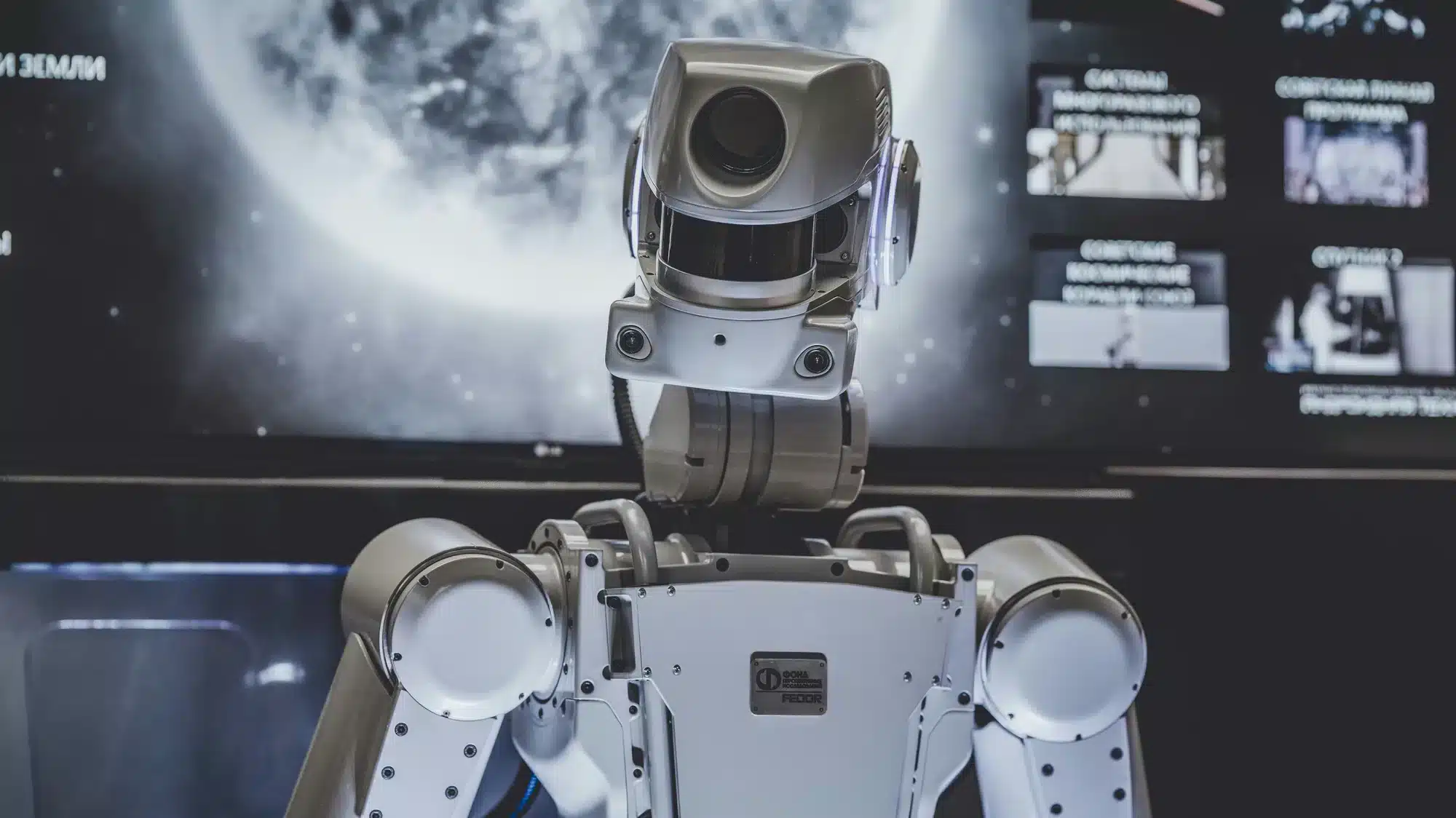 A sleek gray humanoid robot with a camera-like head and articulated arms stands in a high-tech lab with screens in the background.