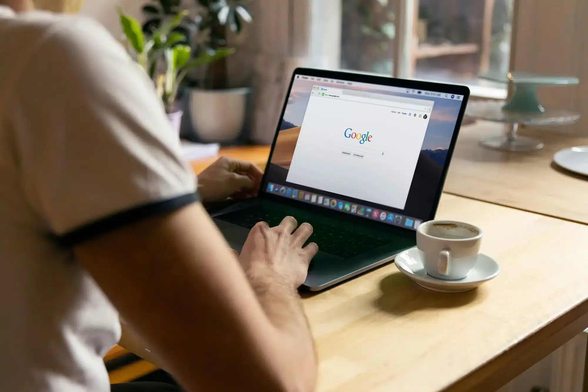 Person typing on a laptop at a wooden table; the screen shows Google, with a cup of coffee on a saucer nearby.