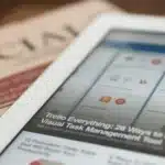 Tablet on a wooden surface displays a Trello dashboard with colorful app tiles; a blurred newspaper sits in the background.