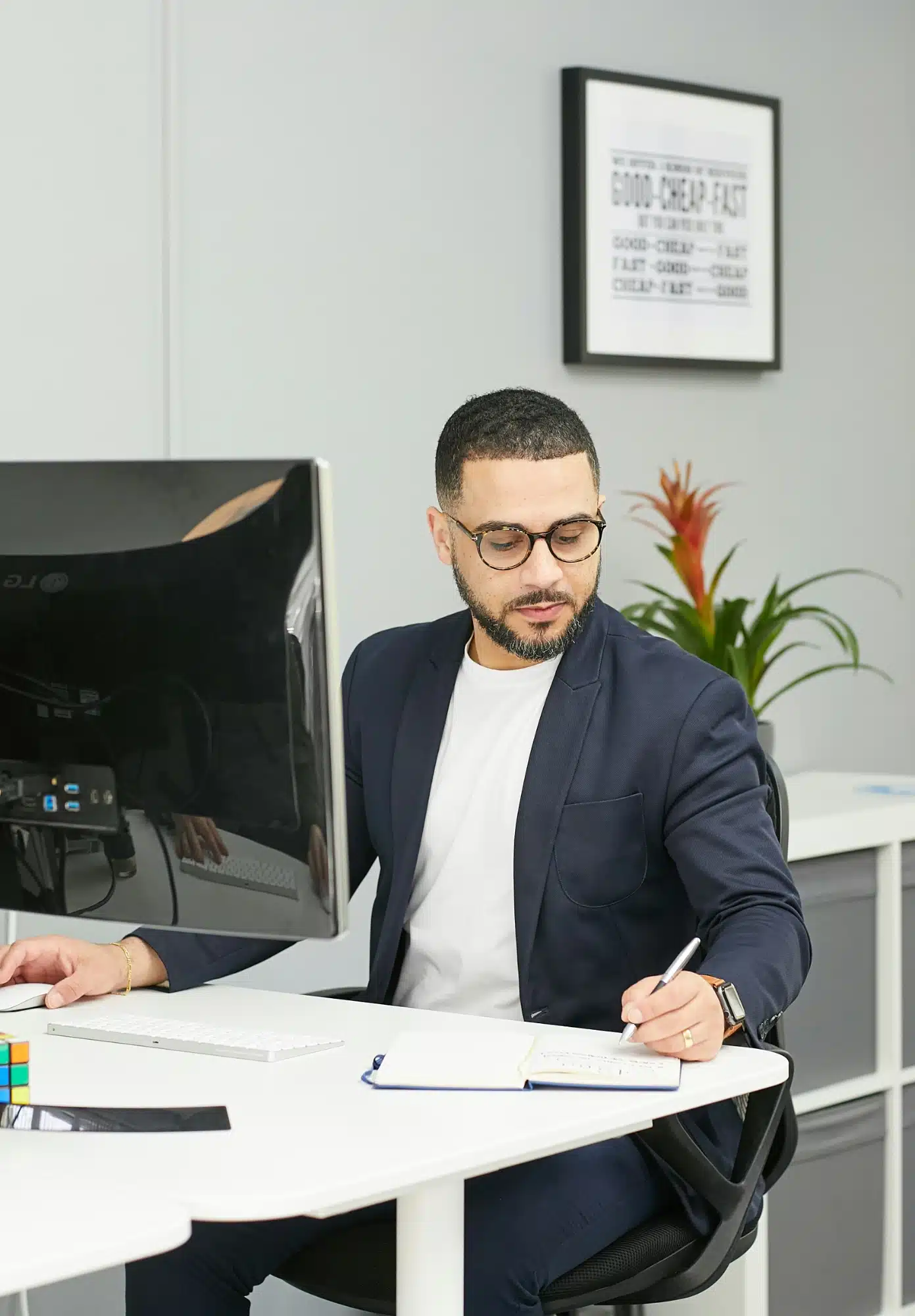 A man with glasses and a short beard, wearing a navy blazer and white shirt, sits at a white desk in a modern office and writes in a notebook.