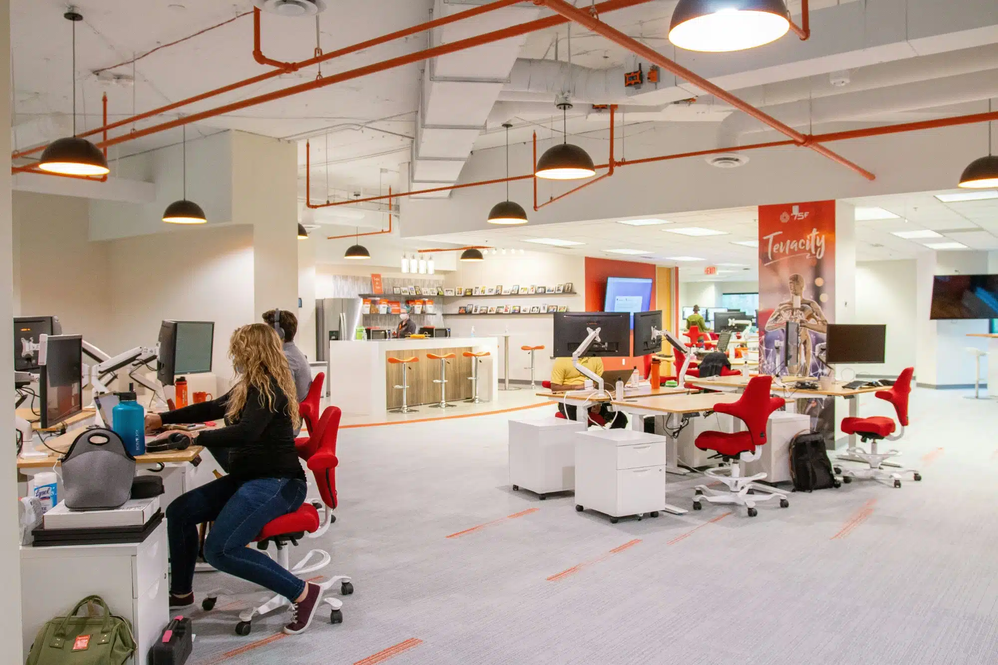 Open-plan office with employees at white desks and red chairs, several computer monitors, orange ceiling pipes, and a bar-height counter in the background.