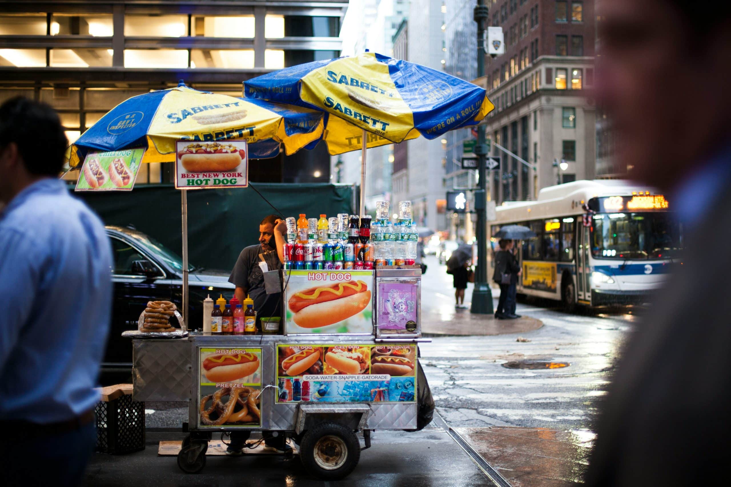 Street hot-dog cart under yellow and blue Sabrett umbrellas, with condiments, bottled drinks, and a hot-dog sign on a rainy city street.