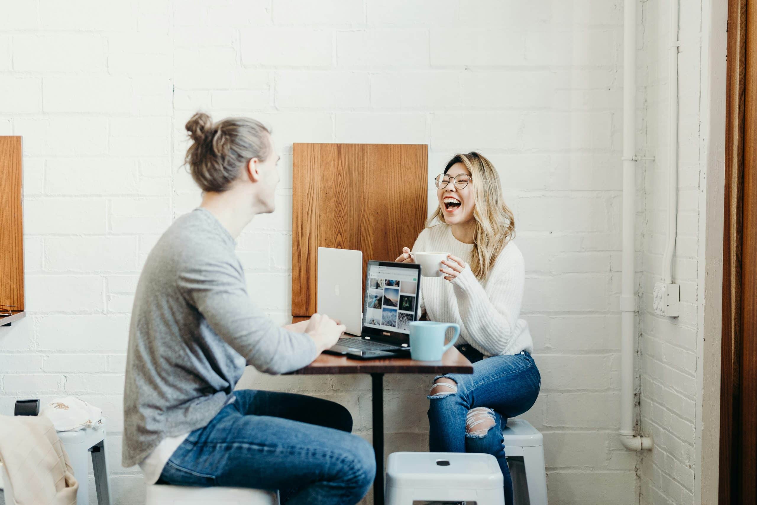 Two friends sit at a small table in a white brick cafe; the woman with glasses laughs, holding a mug beside her laptop.