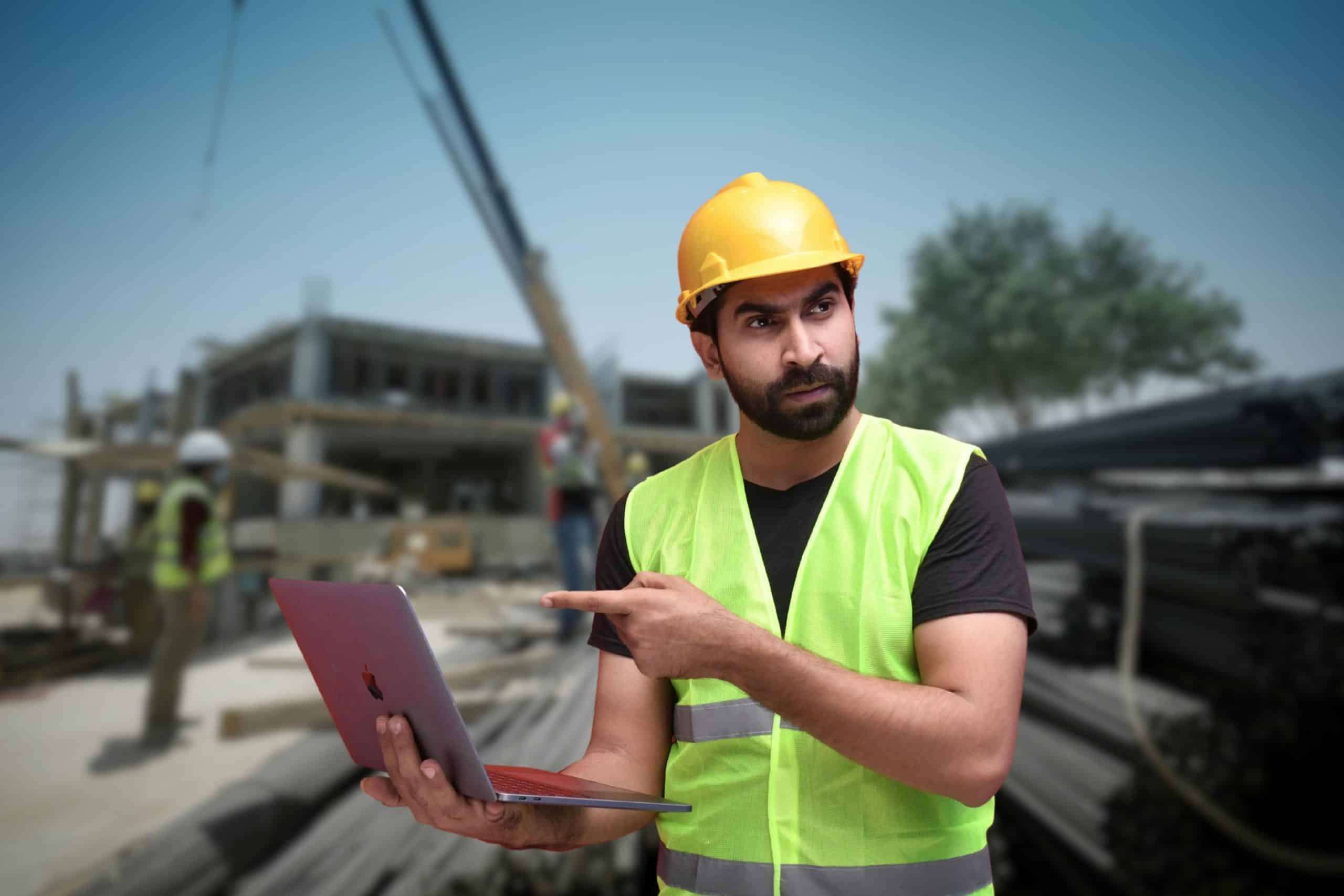 Bearded construction worker in a neon vest and yellow hard hat holds a laptop and points at it on a busy construction site.
