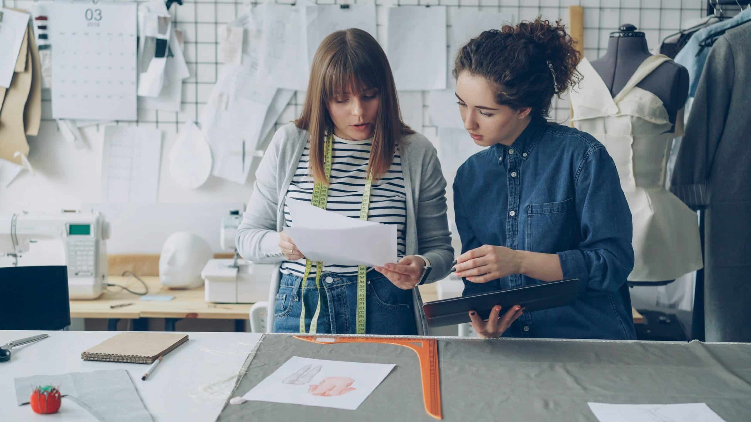 Two fashion design students examine pattern sheets in a sewing studio, with dress forms, fabric, and pattern papers in the background.