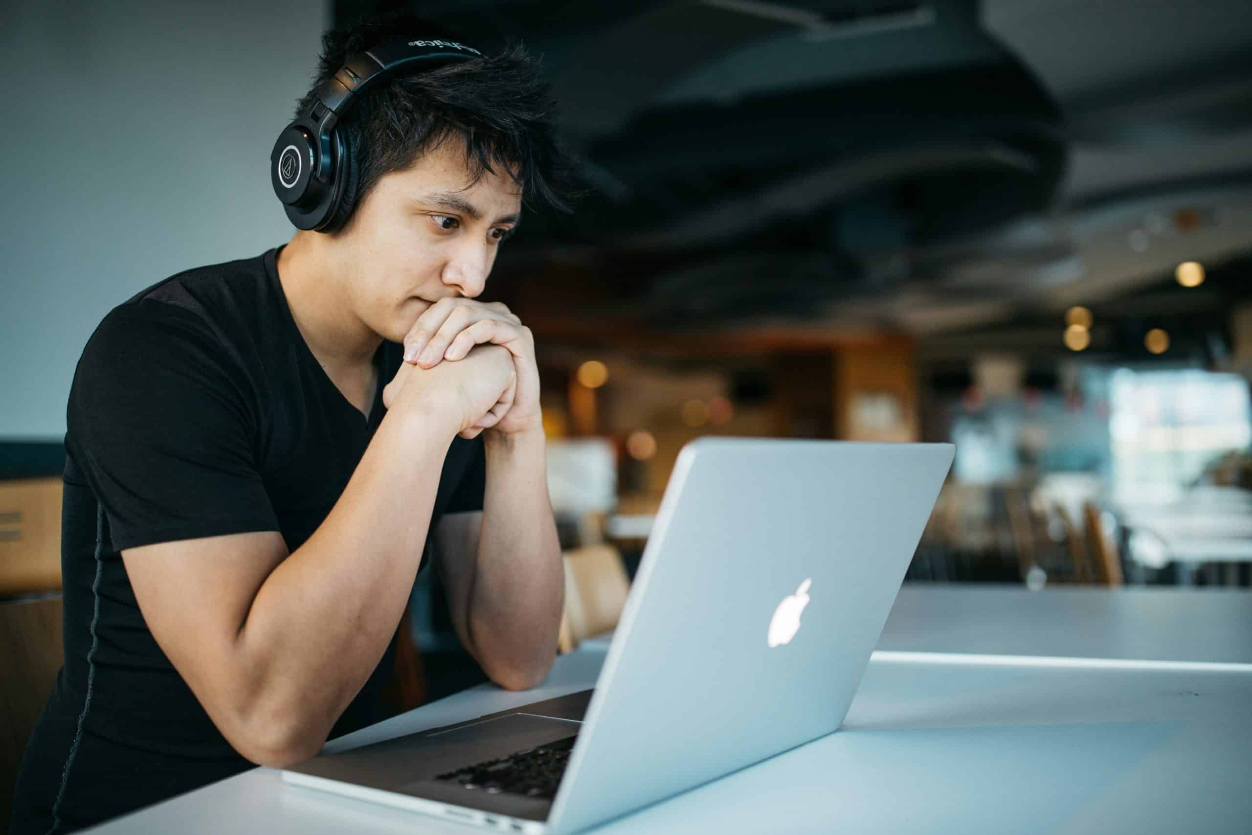 Young man in a black t-shirt wearing over-ear headphones sits at a cafe table, hands clasped, looking at a silver MacBook.