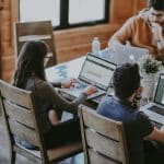 Three people working on laptops at a wooden table, focused on their tasks in a cozy, well-lit space. Photo by Annie Spratt on Unsplash.