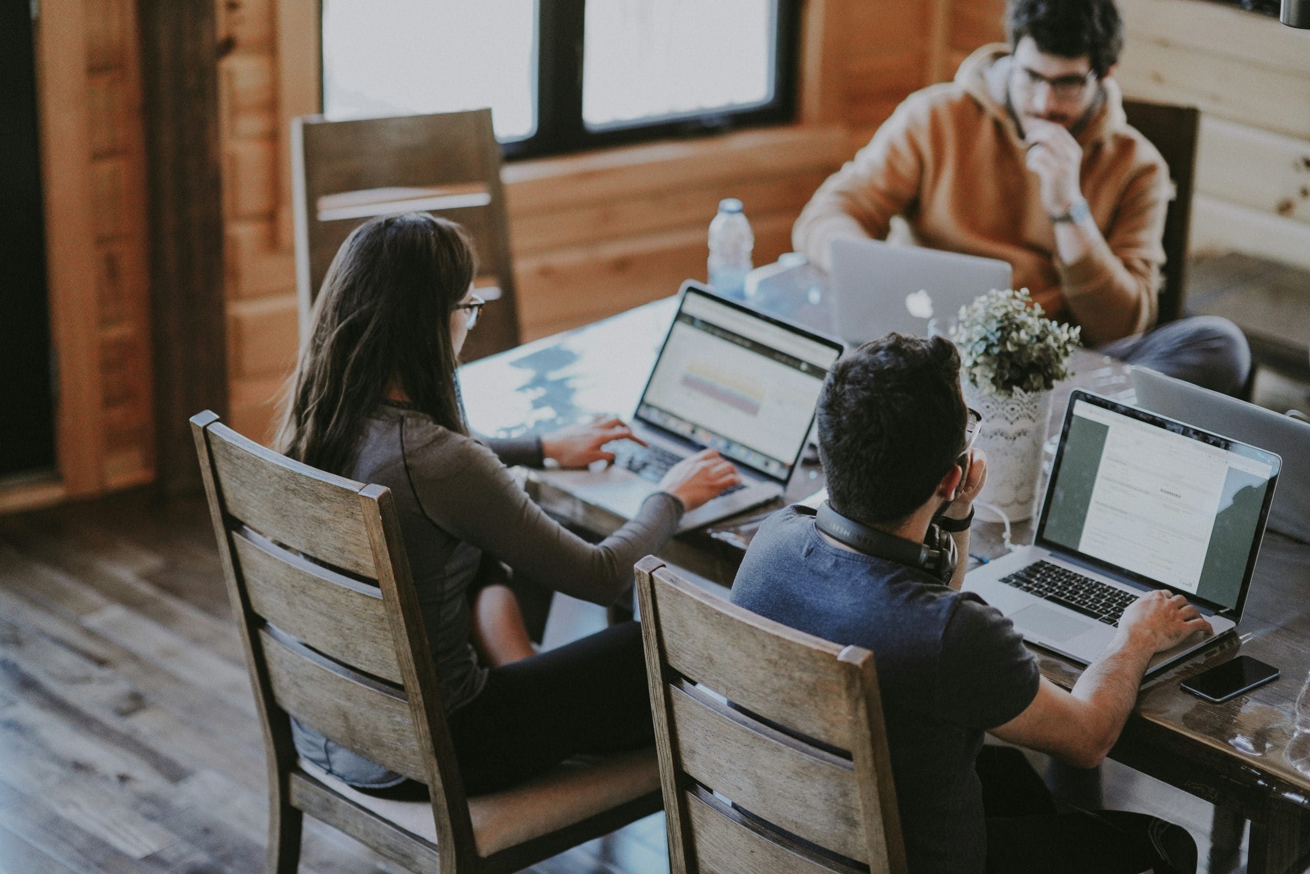 Three people working on laptops at a wooden table, focused on their tasks in a cozy, well-lit space. Photo by Annie Spratt on Unsplash.